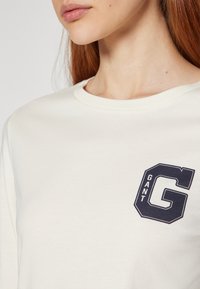 Cremefarbener Sweatshirt mit rundem Halsausschnitt und einem dunkelblau gestickten Logo auf der linken Brust mit der Aufschrift "GANT G." Glatte Baumwolltextur.