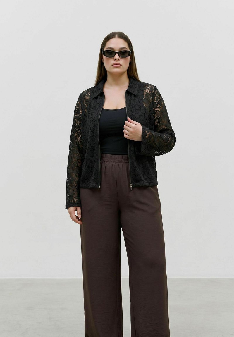 Femme portant une veste en dentelle noire, un haut noir, un pantalon marron à jambes larges et des lunettes de soleil noires, debout devant un fond gris uni.
