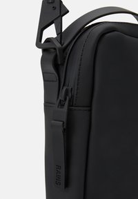 Borsa impermeabile nera con texture in gomma liscia, dotata di un design elegante, tracolla regolabile e un'evidente clip in metallo e dettaglio della cerniera.
