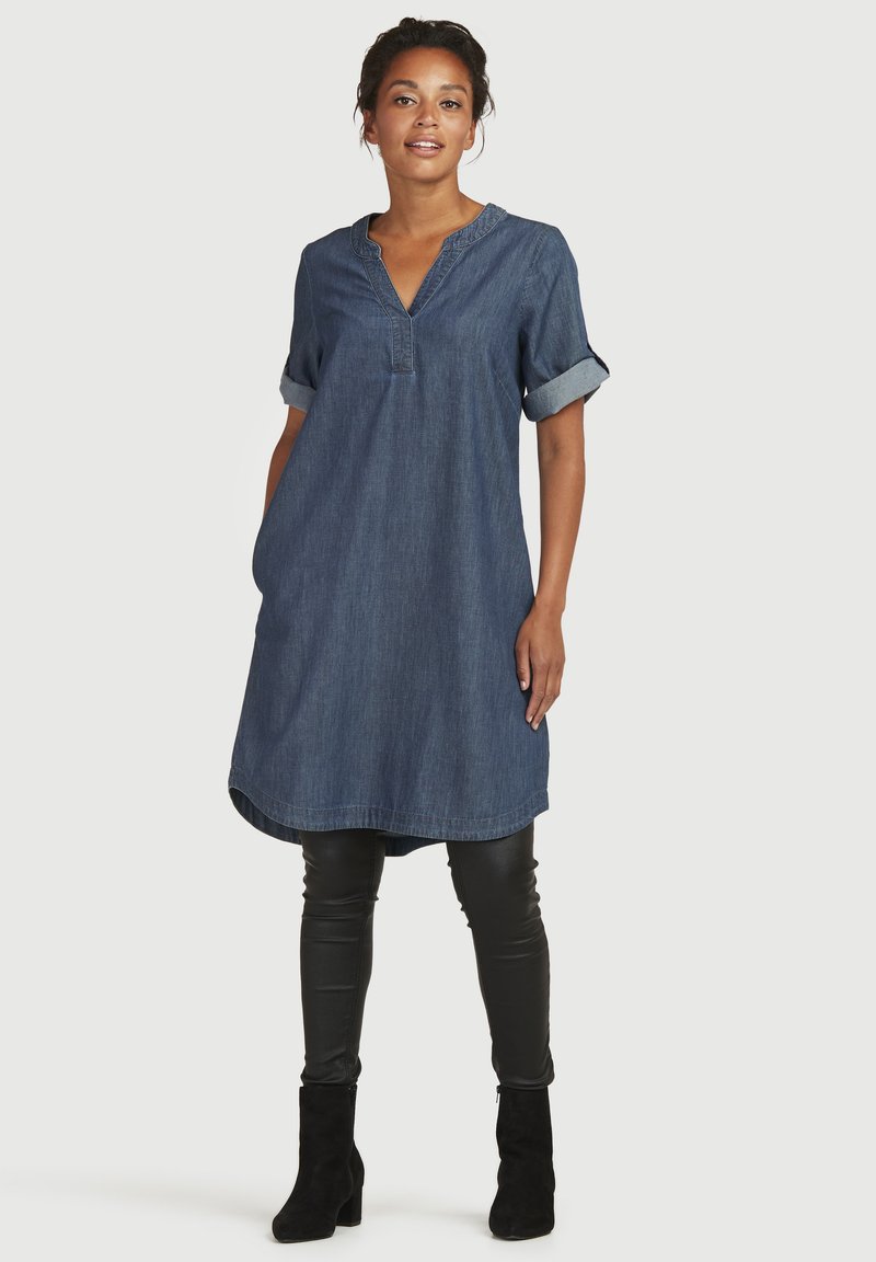 Denim jurk met korte opgerolde mouwen, klassieke V-hals en zijzakken. De zoom is afgerond en valt boven zwarte getailleerde broek en enkellaarsjes.
