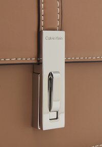 Detail tašky Calvin Klein s kovovou stříbrnou sponou, tělem z opálené kůže a kontrastním prošitím podél okrajů.