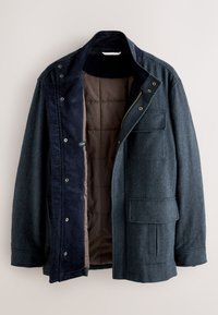 Veste en laine chevrons gris foncé avec doublure matelassée marron, patte intérieure bleu marine, poches à rabat, col montant et boutons pression à l'avant.
