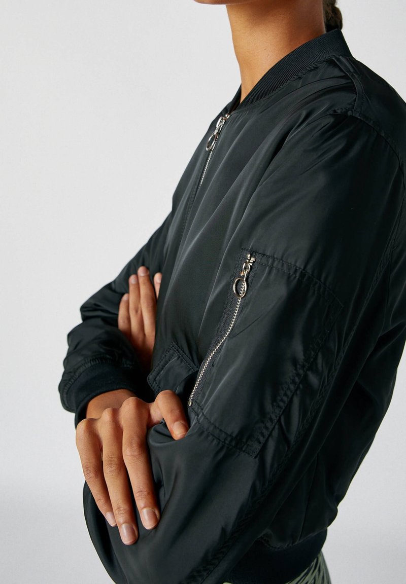 Personne aux bras croisés portant une veste bomber noire avec une poche à fermeture éclair sur la manche, devant un fond uni.