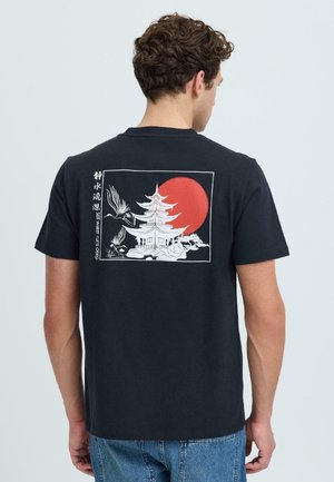 Uomo che indossa una maglietta nera con una pagoda bianca, due gru in volo, sole rosso e testo verticale "L'acqua calma scorre profonda" sul retro.