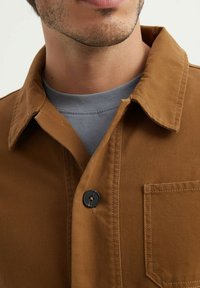 Veste chemise en toile marron avec un col rabattu, un bouton noir unique et une poche poitrine. En dessous, un t-shirt à col rond gris.