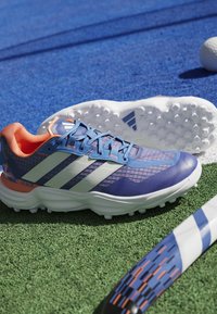 Scarpe sportive blu con strisce bianche, tomaia in mesh testurizzato e dettagli arancioni. Suola in gomma con tacchetti per migliorare la trazione su superfici erbose.