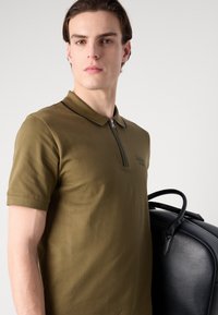 Olive zaļš polo krekls ar īsām piedurknēm, ar rāvējslēdzēju apakšā, un melniem akcenta šuvumiem; modelis tur melnu ādas somu ar teksturētu virsmu.