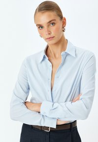 Camicia azzurro chiaro con colletto, maniche lunghe e una tasca sul petto, abbinata a una cintura scura e pantaloni.