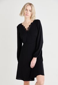 Μαύρο φόρεμα με μακριά μανίκια, V-λαιμόκοψη με δαντέλα από καλοκαιρινή μπορντούρα και άνετη εφαρμογή. Λείο ύφασμα και μήκος μέχρι το γόνατο.