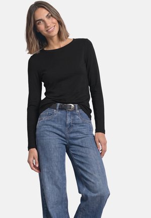 Kvinde med skulderlangt brunt hår iført en sort langærmet trøje og blå jeans med sort bælte, smilende og stående mod en hvid baggrund.