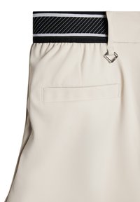 Beżowe spodnie z marszczonym pasem, w którym znajduje się elastyka w czarno-białe paski. Zawierają małą tylnią kieszeń oraz srebrny detal w postaci pętli.