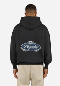 Sort hætte sweatshirt med et stort ovalt grafisk motiv på ryggen, hvor teksten "Pegador" står i hvid, omkranset af blåt og med ternede detaljer.