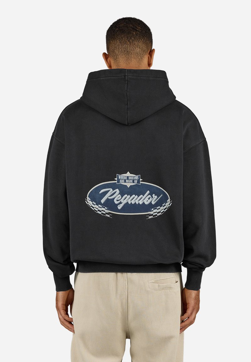 Sort hætte sweatshirt med et stort ovalt grafisk motiv på ryggen, hvor teksten "Pegador" står i hvid, omkranset af blåt og med ternede detaljer.