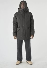 Schwarze Kapuzenjacke mit Knöpfen auf der Vorderseite und zwei großen Taschen. Getragen über dunklen Hosen und hellen Schuhen. Einfaches und funktionales Design.