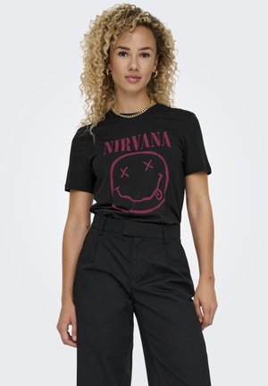 Mujer con cabello rizado rubio que lleva una camiseta negra de la banda Nirvana, aretes de aro dorados, un collar de cadena y pantalones negros de cintura alta, de pie contra un fondo blanco.