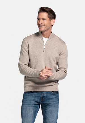 Beige gebreide trui met een ritskraag, geribde manchetten en een tailleband, gecombineerd met blauwe spijkerbroek. Glad textuur en aansluitend ontwerp.
