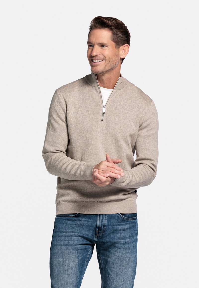 Beige gebreide trui met een ritskraag, geribde manchetten en een tailleband, gecombineerd met blauwe spijkerbroek. Glad textuur en aansluitend ontwerp.