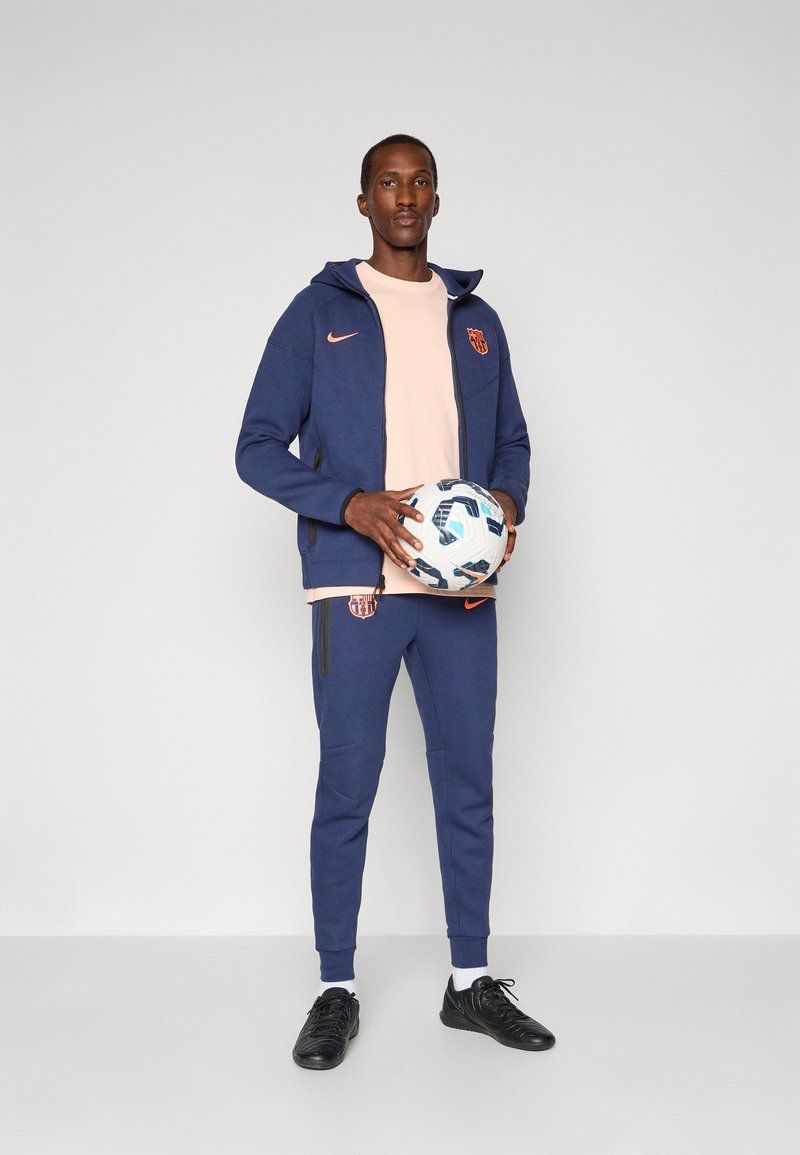 Marineblå hettejakke med glidelås, oransje detaljer og logo, matchende marineblå joggebukser, hvit t-skjorte og svarte treningssko, holder en mønstret fotball.