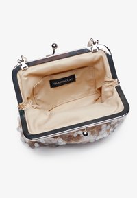 Handtasche mit metallischem Verschluss, ausgestattet mit einem transparenten, mit Perlen besetzten Äußeren sowie einem beigen Stoffinnenraum und einem Etikett im Inneren.