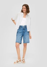 Witte shirt met korte mouwen en opengewerkt detail, gecombineerd met mid-blauwe denim shorts. Model draagt lichtgekleurde sandalen met banden.