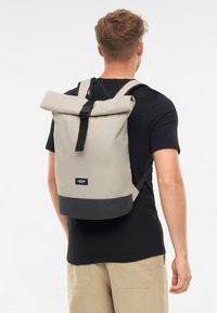 Sac à dos roll-top en gris clair avec un fond noir. Dispose de bretelles réglables et d'une fermeture à boucle à l'avant. Texture de tissu durable.