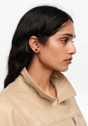 Mujer joven con chaqueta beige y cuello levantado lleva pendiente de botón negro en forma de corazón, mirando hacia la derecha sobre fondo blanco.