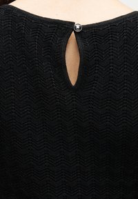 Μαύρο πλεκτό ένδυμα με υφή και μικρό άνοιγμα σε σχήμα δάκρυ, στερεωμένο στην κορυφή με ένα στρογγυλό κουμπί.