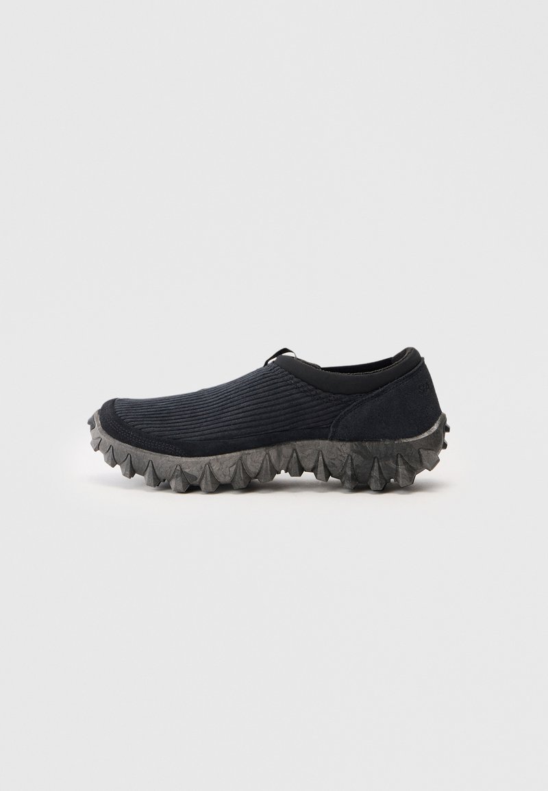 Melnas slip-on kurpes ar ribotu audumu augšdaļu un izturīgu, spikes pelēku zoli, attēlotas profila skatījumā uz vienkrāsaina fona.