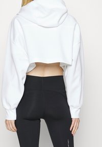 Sudadera blanca corta con capucha, mangas oversize y un corte en la parte posterior, combinada con leggings negros de talle alto. Textura de tela suave.