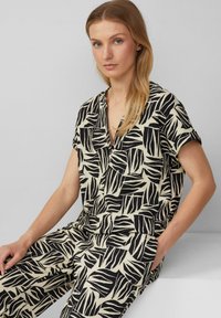 Zwarte en crèmekleurige jumpsuit met bladpatroon, korte mouwen, kraagloos ontwerp, elastische taille en zijzakken. Gestructureerde stof.