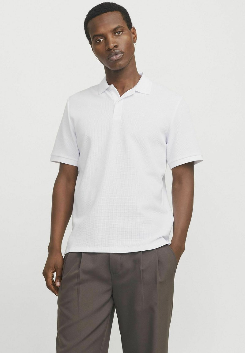 Polo-shirt blanc avec un tissu texturé, manches courtes et une patte de boutonnage à trois boutons. Associé à un pantalon marron ajusté.
