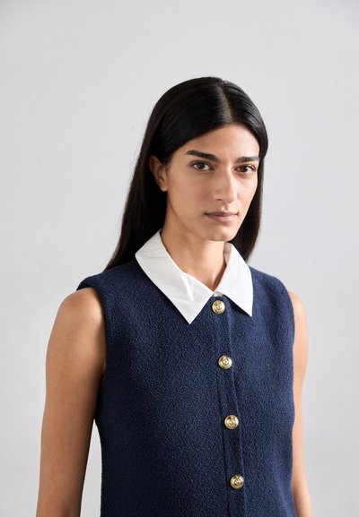 Robe sans manches texturée bleu marine avec un col blanc, présentant des détails de boutons dorés sur le devant. Tissu lisse et ajusté.