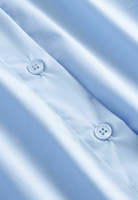 Tissu bleu clair avec deux boutons ronds cousus sur une couture centrale, texture lisse avec des plis doux.