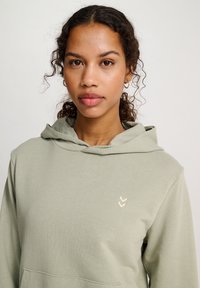 Hoodie vert clair en tissu doux, avec une poche kangourou et un petit logo doré sur la poitrine.
