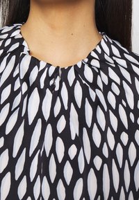 Gros plan d'un chemisier à motifs noir et blanc avec un petit bouton près du col, porté par une personne aux longs cheveux foncés.