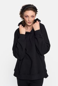 Czarna, oversize bluza z kapturem, wykonana z miękkiego materiału, z regulowanym kapturem i ściągaczami przy rękawach. Sylwetka jest luźna z warstwowym wykończeniem dołu.
