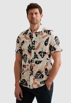 Mann trägt ein kurzärmeliges Hemd mit Knöpfen und schwarzen sowie braunen abstrakten Blattmustern auf beigem Hintergrund, steht mit einer Hand in der Tasche.