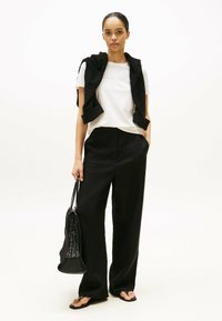 Camiseta blanca con texto bordado, suéter negro de punto drapeado sobre los hombros, pantalones anchos negros y una bolsa de malla negra.