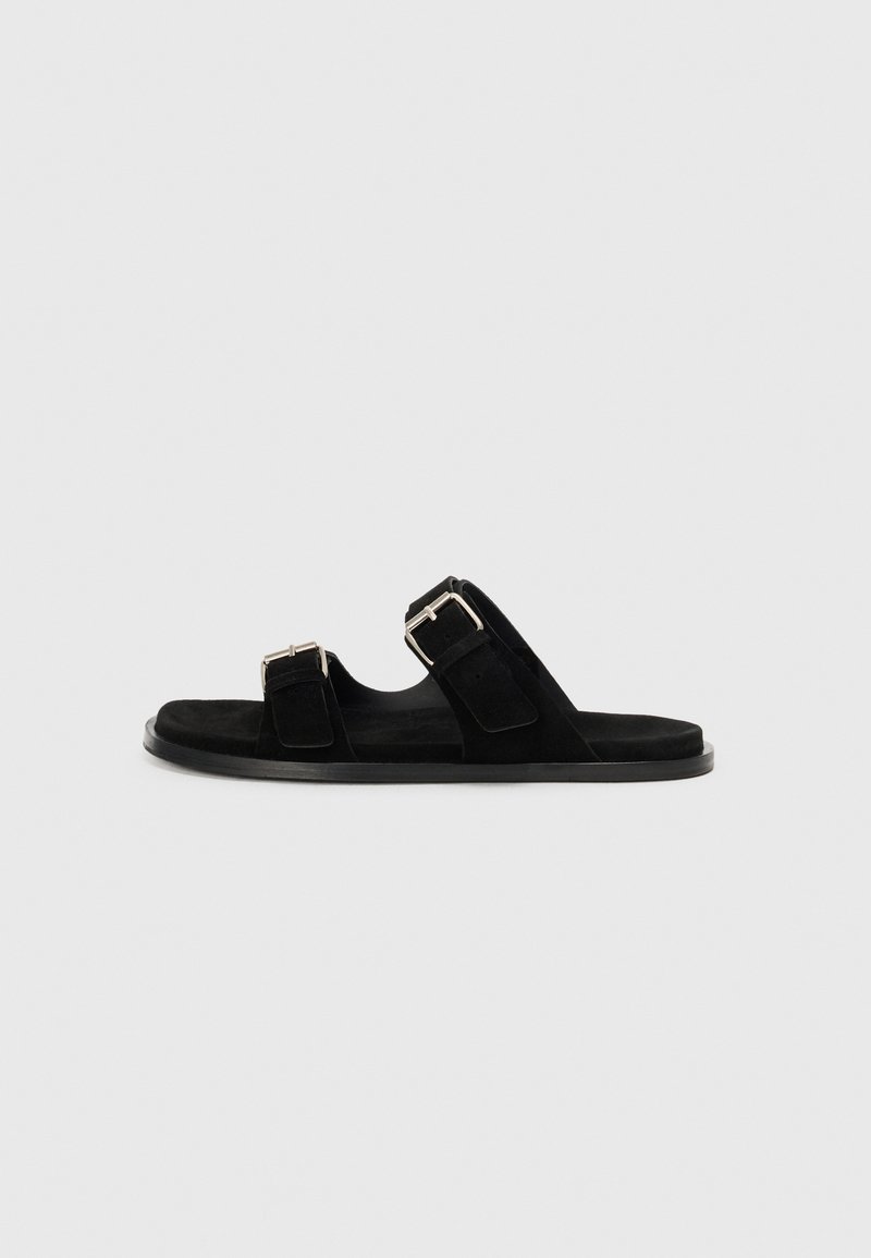 Sandali neri in suede con due cinturini regolabili, fibbie argentate, suole piatte e design aperto, che offrono un'estetica minimalista.