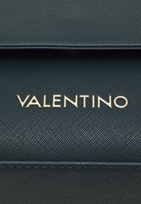 Tamsiai žalia tekstūruota odinė rankinė su auksiniu "VALENTINO" logotipu, pasižyminti elegantišku dizainu ir subtiliomis siūlėmis palei kraštus.