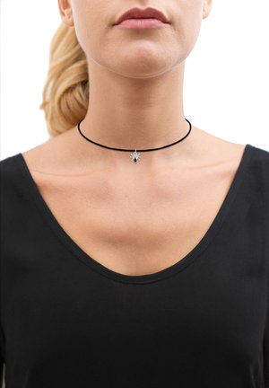 Črna choker ogrlica s srebrnim obeskom v obliki pajka, ki ima osrednjo črno drago, nosi se z črnim V-izrezom proti beli podlagi.