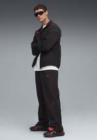 Černá bunda a kalhoty s červenými akcenty, bílé tričko a tmavé sportovní tenisky se barevným designem. Černé oversized sluneční brýle.