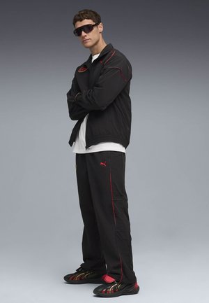 Svart jacka och byxor med röda detaljer, en vit T-shirt och mörka sportiga sneakers med en flerfärgad design. Stora svarta solglasögon.