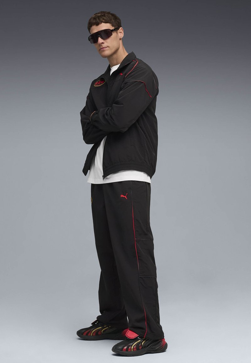 Černá bunda a kalhoty s červenými akcenty, bílé tričko a tmavé sportovní tenisky se barevným designem. Černé oversized sluneční brýle.