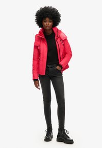 Pink isoleret jakke med høj krave og lynlås, parret med en sort sweater og skinny jeans samt sorte snørebånds støvler.