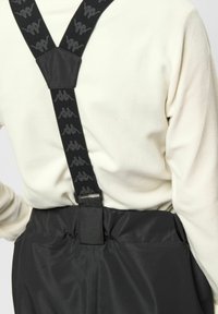 Personne portant des bretelles noires avec un logo répété sur une chemise blanche à manches longues et un pantalon noir, vue de dos.