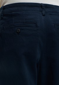 Pantalon bleu foncé montrant la poche arrière avec un bouton et des passants de ceinture, porté avec une chemise blanche rentrée.
