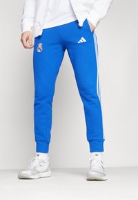 Blue sweatpants featuring white Adidas stripes and Real Madrid logo, crafted from soft fabric with a tapered fit and elastic cuffs.