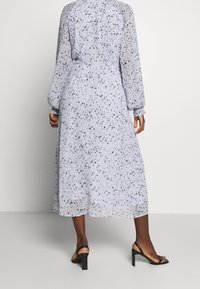 Helles blaues, langärmliges Kleid aus transparenter Stoff, mit einem schwarz gesprenkelten Muster, tailliertem Bund und knöchellanger Rocklänge. Getragen mit schwarzen Absätzen.