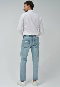 Vaqueros de color azul claro con corte recto, que presentan detalles desgastados en los bolsillos traseros y un sutil pespunte en contraste, combinados con una camisa blanca.